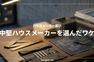 デスクライトに照らされたモダンな書斎の机。アンティーク調の金属製メジャーや、丸められた青い建築図面、プレカット木材、コンクリートブロック、ペンケースが並ぶスタイリッシュな作業風景。「ハウスメーカー選び 中堅ハウスメーカーを選んだワケ」の文字タイトル入り。