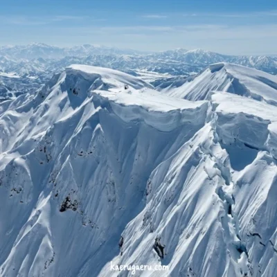 青空の下、巨大な雪庇が形成された険しい雪山の稜線と、遠くに連なる山々。
